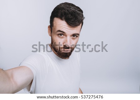 Handsome young man in white shirt doing selfie. Attractive guy in white shirt with beard is making a funny face mocking of girls with pumped lips. Isolated on white background