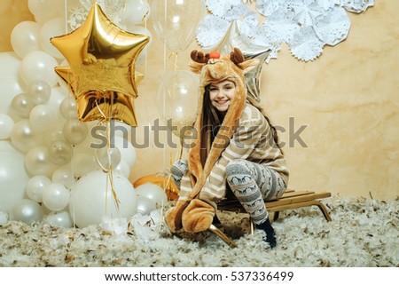 small christmas cute child or smiling girl in winter pants, sweater and animal hat with wooden sledge near silver and golden stars on white balloons and decorative new year snowflakes background