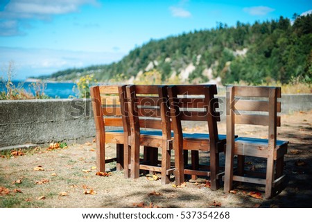 A row of faded, old wooden chairs on a light dirt shore sets a serene scene amidst a lake on a sunny day with lush green hills in the background.