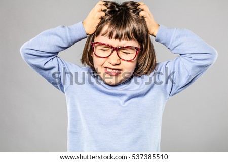 irritated 6-year old young girl with eyeglasses pulling out her hair for itchy allergies or lice or scratching her head for nervous disagreement, grey background 