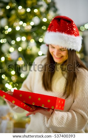Boxing day. Nice girl the brunette opening a box with the Christmas present. On her face joy. Behind the girl beautifully decorated Christmas tree.