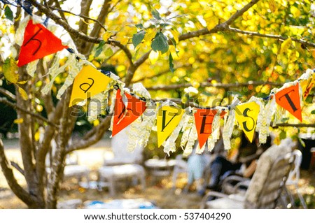 Orange and yellow harvest sign strung over frame with golden light, soft focus fall trees, leaves, dappled light and natural party scene in soft focus background at an orchard.