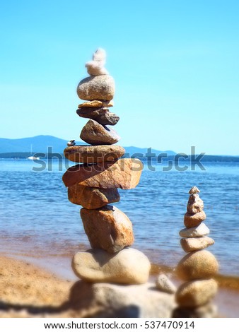 Two rock cairns on beach overlooking calm blue lake, mountains, and white sailboat - soft focus