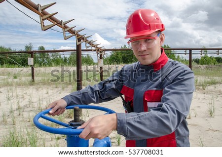Worker near wellhead valve holding radio and wearing red helmet in the oilfield. Pipeline background.  Oil and gas concept.