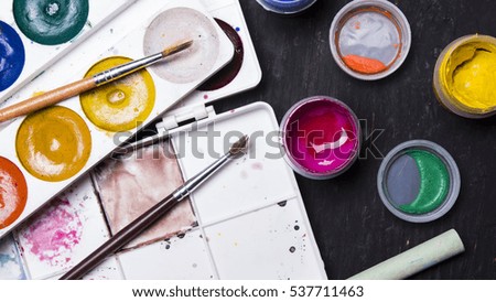 Bright multi-colored paint, pastels, crayons, pencils, brushes on a black wooden background. Set for the artist. Education.