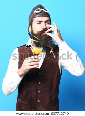 handsome bearded aviator man with long beard and mustache on satisfied face holding glass of alcoholic beverage in vintage suede leather waistcoat with hat and glasses on blue studio background