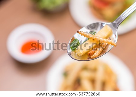 Hand holding wood worm fried Insects with Potatoes in a spoon and there is hamburger, french fries on wooden table in the background. Closeup shot, Select focus.