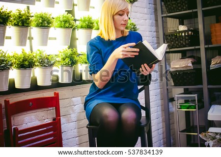 blonde woman in a dress sits on a chair with a book in hand, against the backdrop of houseplants. engaged in reading