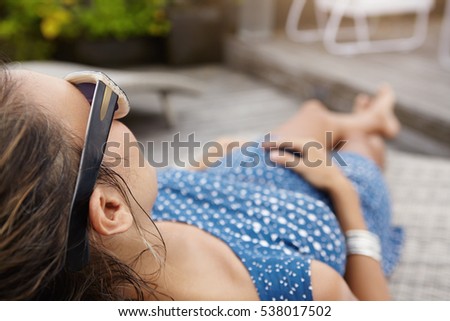 Rear view of beautiful young woman expecting baby lying on sunlounger while spending nice time at spa resort. Pregnant female in shades and blue dress sunbathing during vacations in tropical country