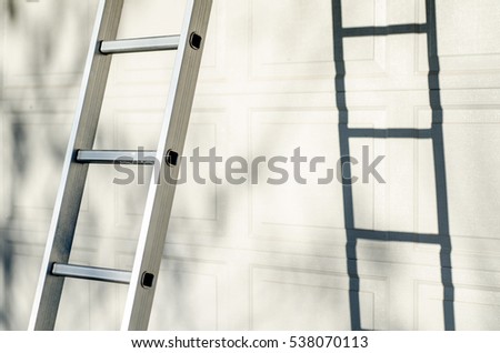 Aluminum ladder leaning against white garage door, with strong shadow and warm natural sun light Royalty-Free Stock Photo #538070113