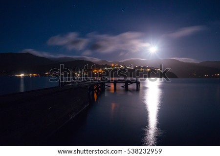 Beautiful moonrise at a lake in Akaroa New Zealand. The moon rises above the mountains while clouds are moving across the horizon above a  old jetty. This is a long exposure shot.