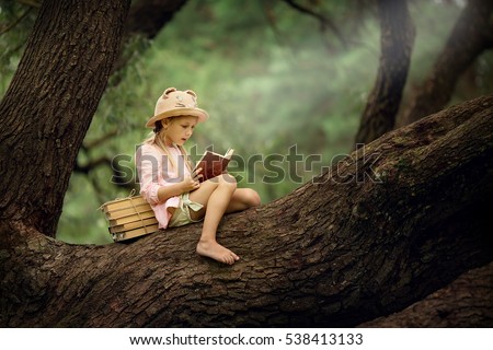 A pretty little blonde girl in a straw hat reading a book on a large spreading tree. Children and science.