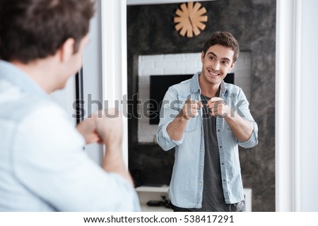 Smiling young man pointing fingers at his mirror reflection while standing at home Royalty-Free Stock Photo #538417291
