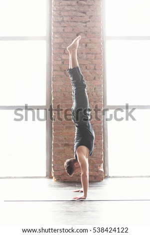 Sport and yoga. Strong young athletic man doing exercise -Handstand against an urban background with picture window and red brick wall on black wooden floor, full length