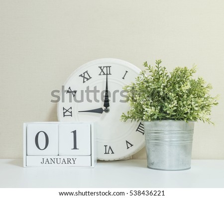 White wooden calendar with black 1 january word with clock and plant on white wood desk and cream wallpaper textured background , selective focus at the calendar