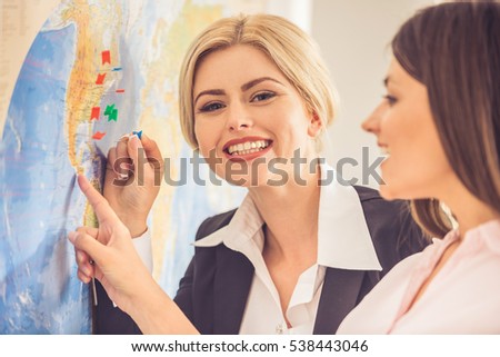 Beautiful female travel agent and her client are standing near map with destination signs on it, choosing place and smiling