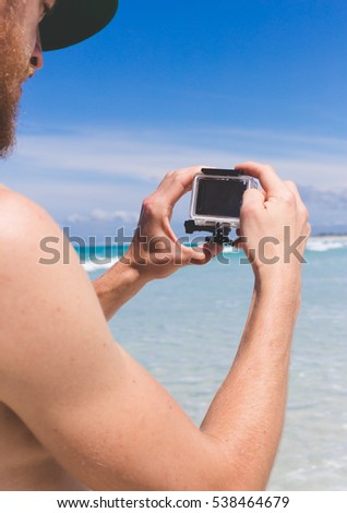 view on hands holding small modern action camera filming on beach with white sand and turquoise water in backdrop, camera has black screen and waterproof housing