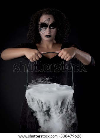 Teen girl dressed up for halloween like a witch and is making a spell potion in her cauldron; on dark black background 