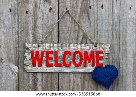 Red welcome sign with blue country fabric heart hanging by rope on antique rustic wood door; Valentine's Day background
