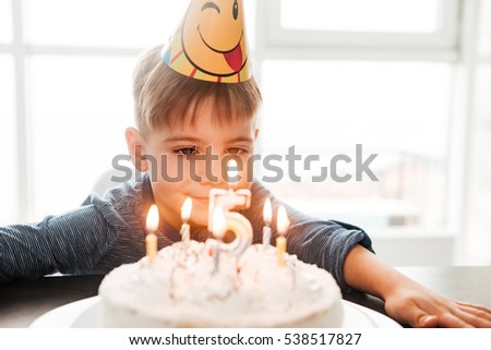 Image of cheerful birthday boy sitting in kitchen near cake while smiling. Look at cake