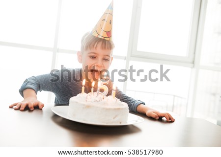 Image of little happy birthday boy sitting in kitchen near cake while smiling. Look at cake