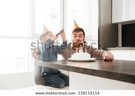 Picture of funny bearded young father sitting near birtday cake with his little son.