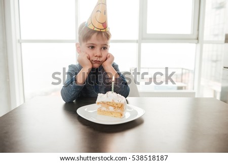 Image of little sad birthday boy sitting in kitchen near cake alone. Look aside.