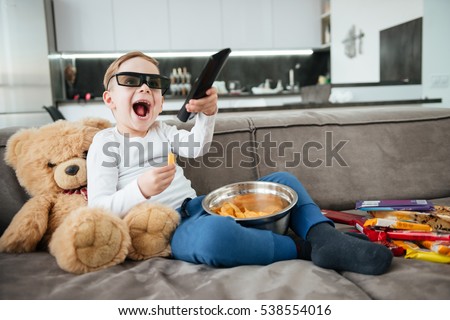 Picture of happy boy on sofa with teddy bear at home watching TV with 3d glasses while eating chips. Holding remote control.
