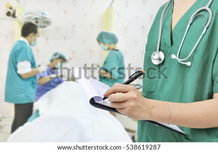 Doctor in the green shirt with stethoscope writing the patient chart on blur operating room background.