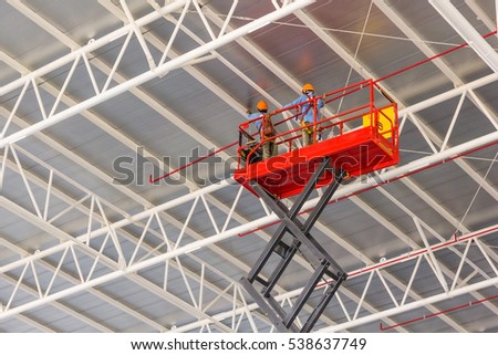 Scissor lift platform with hydraulic system elevated towards a factory roof with construction workers, Mobile aerial work platform Royalty-Free Stock Photo #538637749