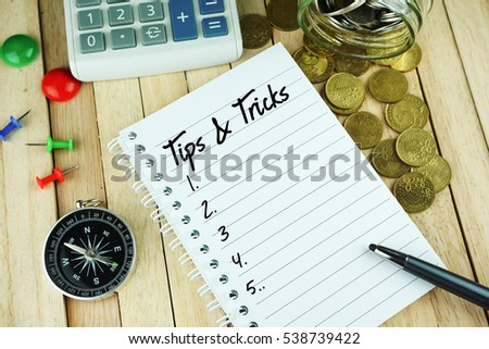 Top view of notebook written with TIPS AND TRICKS. Compass, note pins, calculator, coins, pen and notebook on wooden background. Business Conceptual.