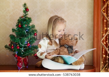 a little girl sitting at home near the Christmas tree and reads a children's winter wonderland, sitting next to a teddy bear