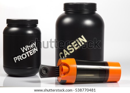 Whey protein black jar and big Casein protein black jar. Orange shaker lying near with opened cap. Close up image. White background. Shadows. Royalty-Free Stock Photo #538770481