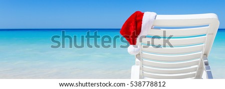 Santa Claus Hat on sunbed near tropical calm beach with turquoise caribbean sea water and white sand. Christmas vacation celebration Royalty-Free Stock Photo #538778812