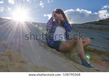 Young beautiful woman backpacker traveling in the desert and making pictures with the vintage camera. Sandy dunes and blue sky on sunny summer day. Travel, adventure, freedom concept.