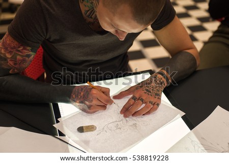 Young male tattooist with tattoos on hands creating sketch in parlor