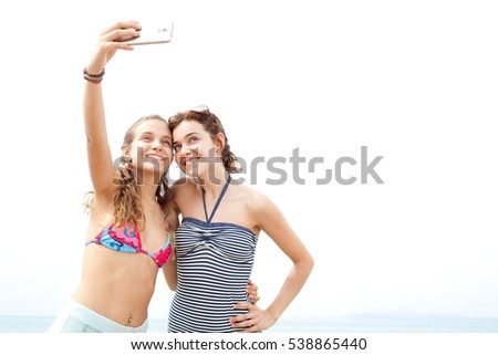 Portrait of sisters on a summer beach holiday joyfully smiling with heads together posing for a selfie photo, holding a smart phone up against sunny sky, outdoors. Travel technology lifestyle.