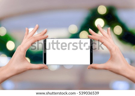 female holding a smartphone isolated blank screen with two hands on Abstract blur background of warm light bokeh on green Christmas pine roping, ready for snap a picture