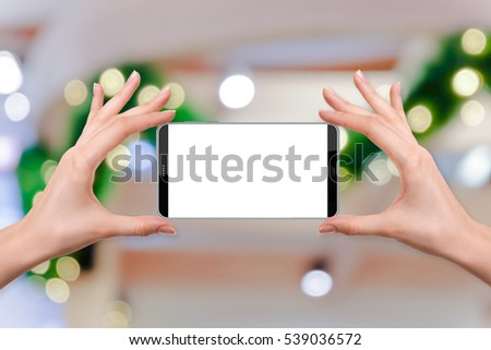 female holding a smartphone isolated blank screen with two hands on Abstract blur background of warm light bokeh on green Christmas pine roping, ready for snap a picture