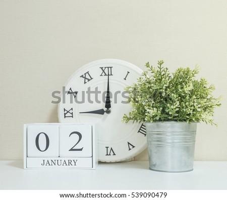 White wooden calendar with black 2 january word with clock and plant on white wood desk and cream wallpaper textured background , selective focus at the calendar