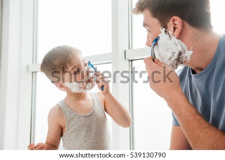 Photo of young happy father and son are applying shaving foam on their faces and smiling while shaving in bathroom Royalty-Free Stock Photo #539130790
