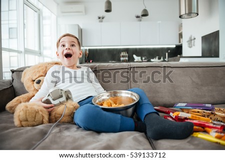Picture of happy boy on sofa with teddy bear at home playing games by console while eating chips. Holding joystick.