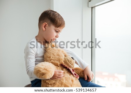 Picture of sad boy near window with teddy bear waiting for parents at home. Look aside.