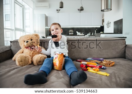 Picture of cheerful boy sitting on sofa with teddy bear at home and watching TV while eating chips. Holding remote control.