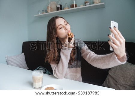 Picture of cute lady sitting in cafe and making selfie with cookie. Look at phone.