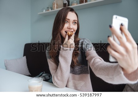 Image of crazy lady sitting in cafe and making selfie with cookie. Look at phone.