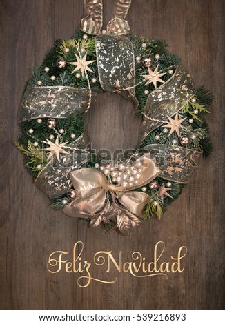 Christmas wreath with golden decorations on wooden door. Text on the picture means "Merry Christmas" in Spanish. This image is toned.