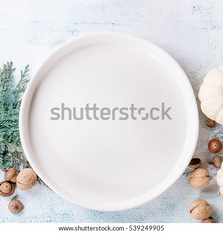 Holiday table decoration with white decorative pumpkins, thuja branches, walnuts, acorns and empty dinner plate over white wooden background. Top view. Square image
