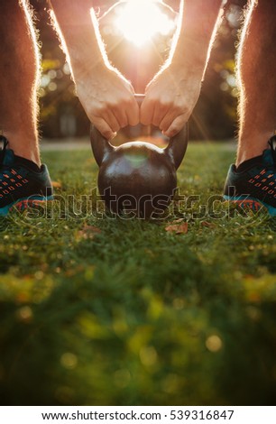 Cropped shot of young man doing kettlebell workout, focus on hands holding kettle bell on grass.