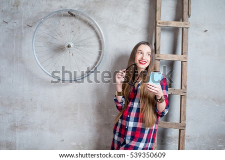 Young woman with phone in checkered shirt standing in the old room with ladder and wheel on the gray textured wall background